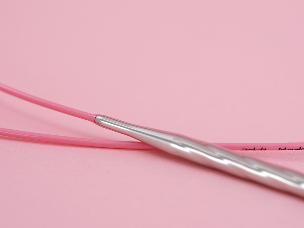 A close-up of a pink knitting needle with a textured silver metal tip resting on a solid pink background. The join between the cable and tip is very smooth.